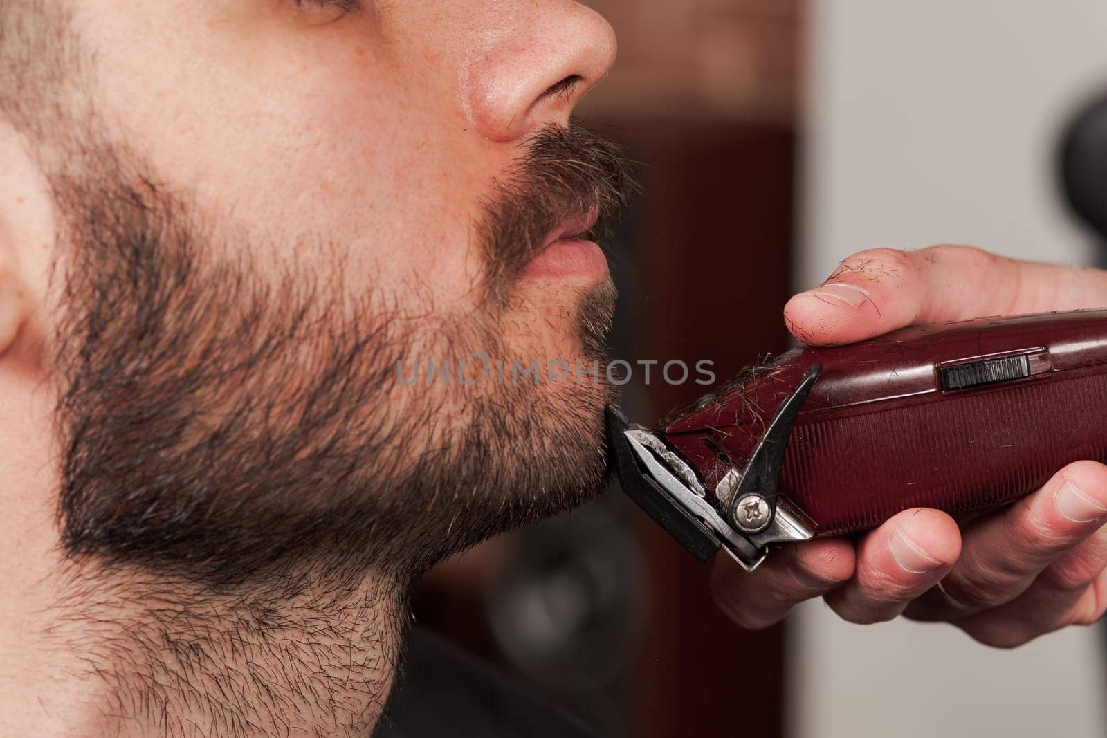 Hipster client visiting barber shop. The hands of young barber making the cut of beard