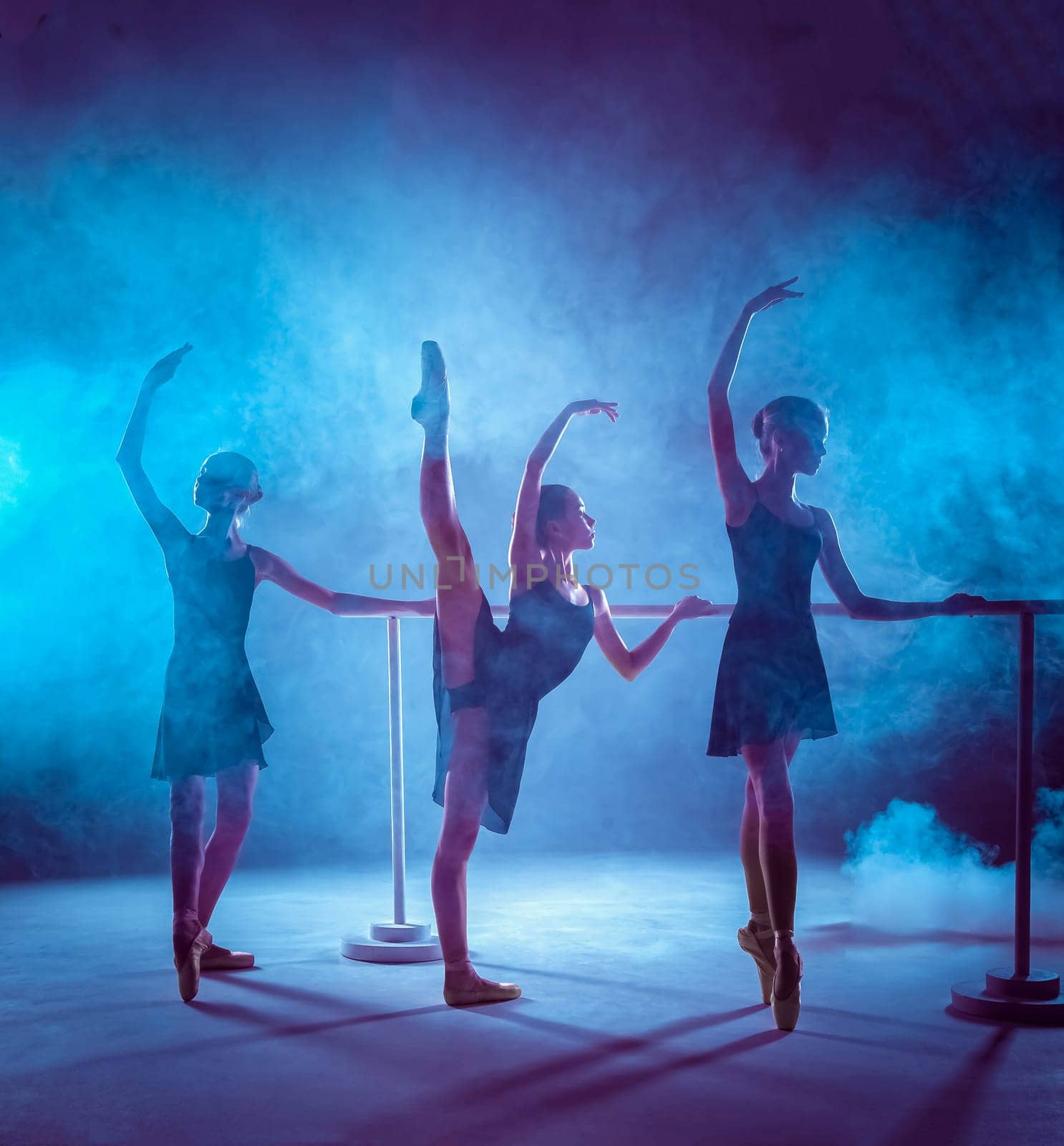 The silhouettes of young ballerinas stretching on the bar on blue smoke background