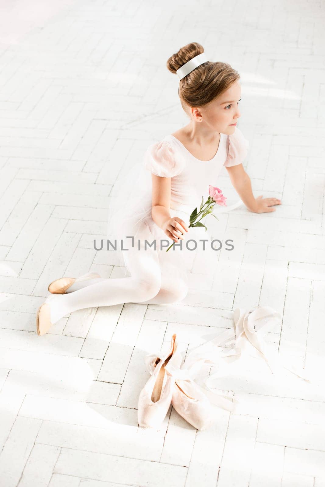 The little balerina in white tutu in class at the ballet school sitting on the wooden floor with flowers