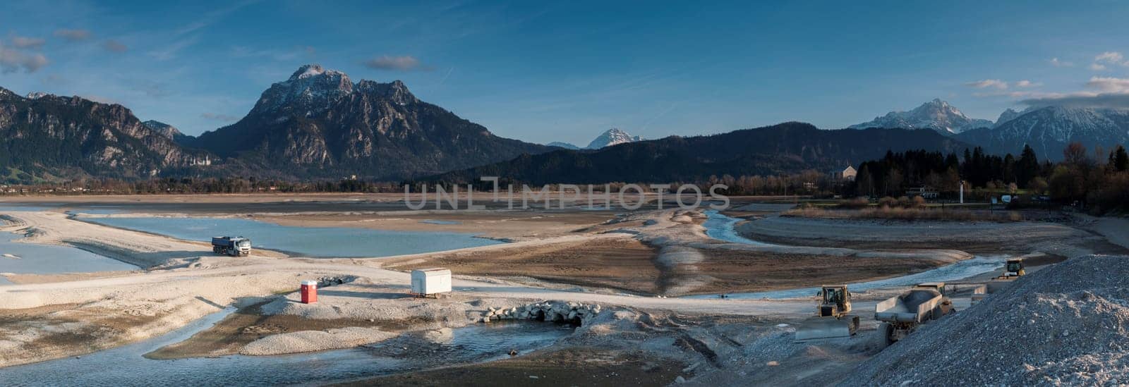 Panoramic view of the drained Forggensee near Füsssen.. The Forggensee,also stored Roßhaupten,is a reservoir crossed by the Lech near Füssen in the Königswinkel in the Bavarian Allgäu. With the meltwater from the surrounding mountains,sedimentary rock and debris settle in the Forggensee,which is collected and transported in winter with drained water,by excavators,bulldozers and other heavy equipment.