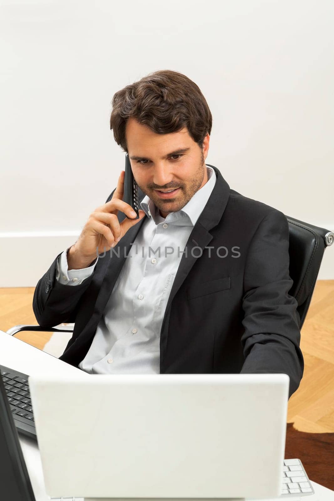 Successful businessman sits in the office and makes phone calls at his computer workplace in black suit