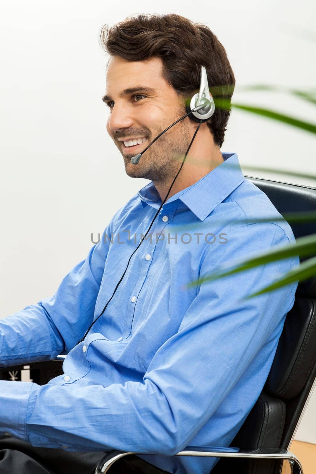 Attractive young man wearing headset in office at customer service operator phone service cheerful with blue shirt