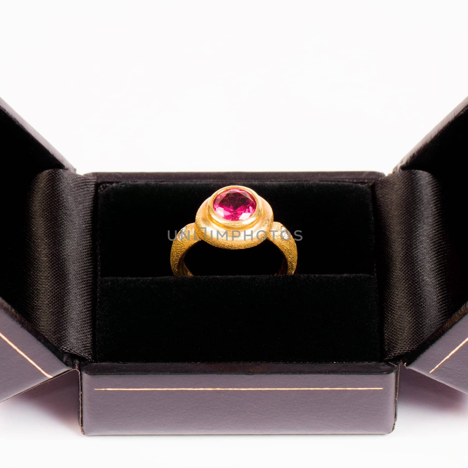 Golden ring with pink gem is in black jewelry box on white background.