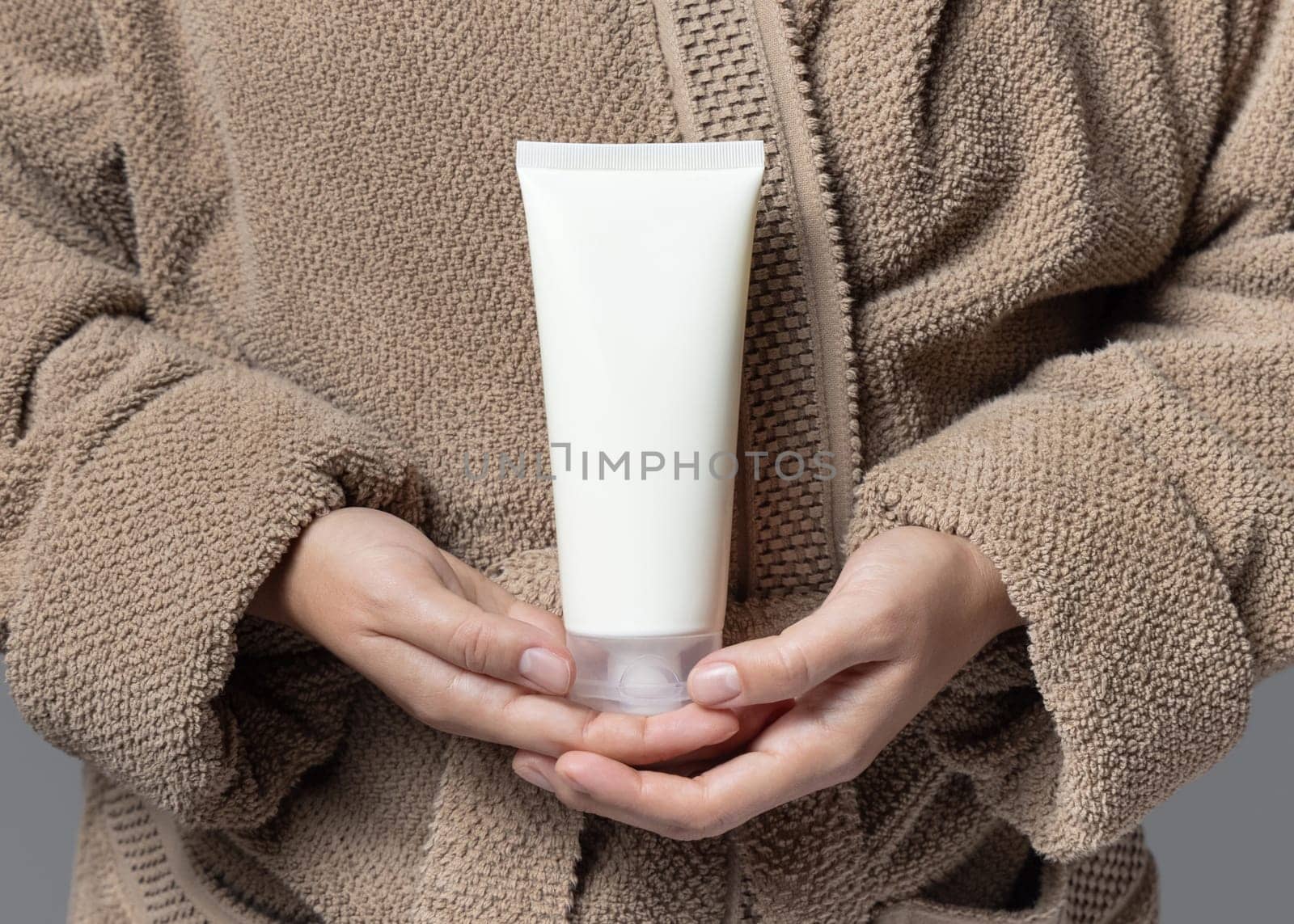 Close up of woman in light brown bathrobe with white blank cream tube in hands, mockup. Hands with natural manicure holding squeeze cosmetic tube - lotion or shampoo