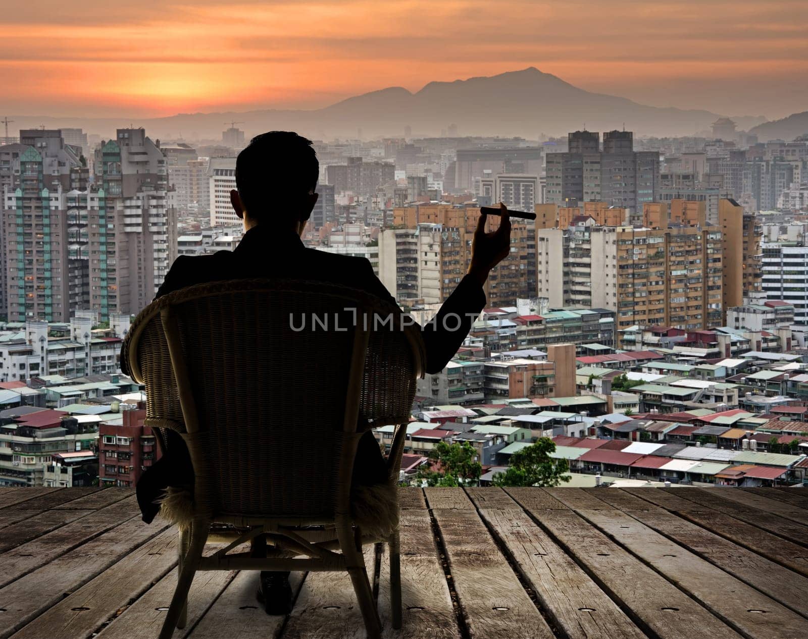 Silhouette of businessman sit on chair and hold a cigar and looking at the city in night.