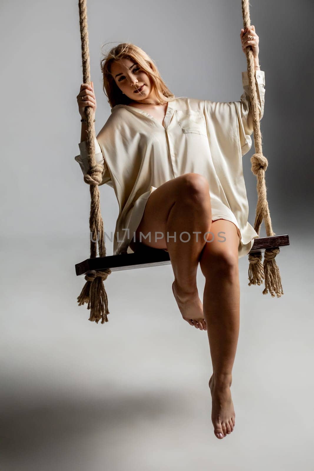 A beautifully captivating woman elegantly poses on a swing, exuding charm and grace in a softly lit artistic space