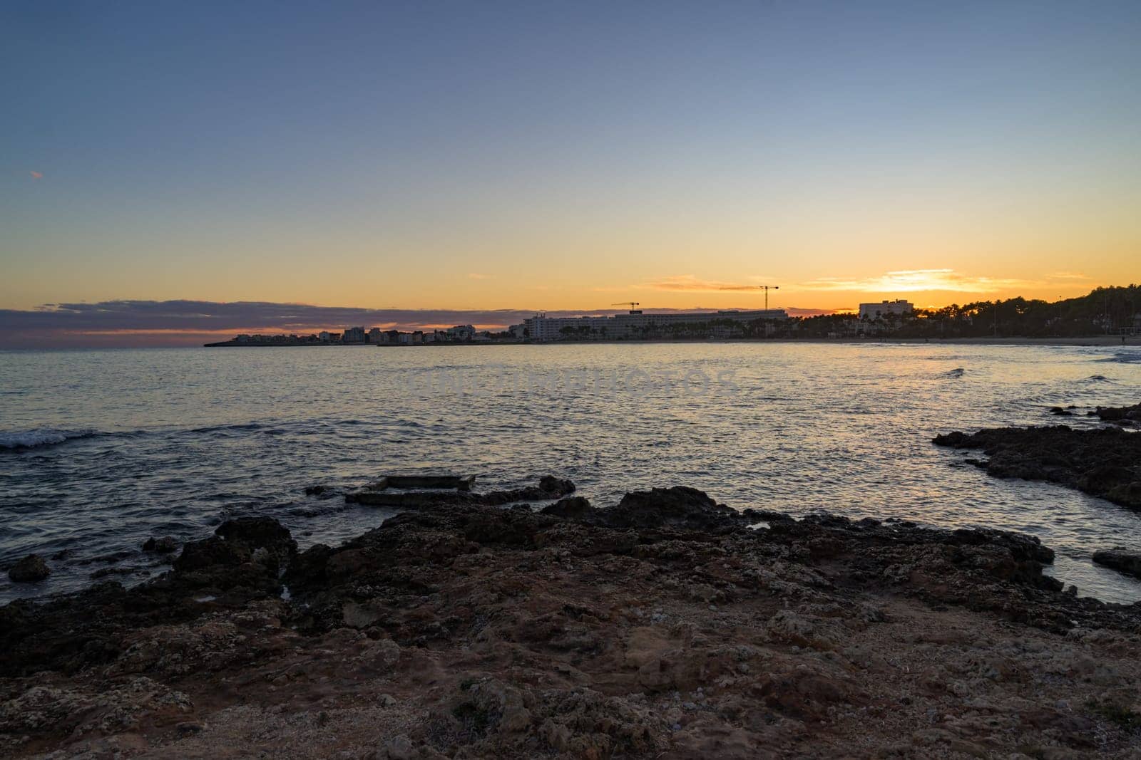 Scenic sunset over the rocky coastline in Mallorca, Spain. The golden light reflects on the calm sea, with buildings and palm trees silhouetted in the background.