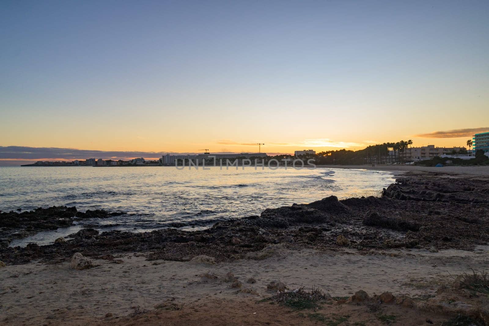 Scenic sunset over the rocky coastline in Mallorca, Spain. The golden light reflects on the calm sea, with buildings and palm trees silhouetted in the background.