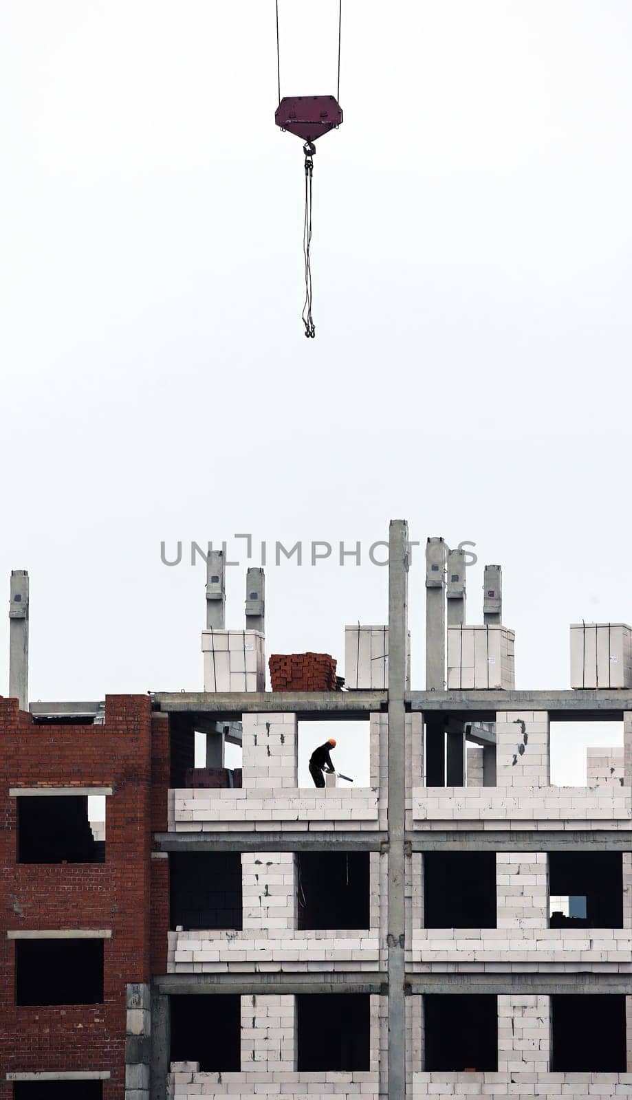 Amidst the backdrop of an overcast sky, construction workers diligently engage in the building of a large structure. A towering crane hovers above, carefully lifting construction materials to the upper levels of the framework. Below, men are seen maneuvering through the partially completed walls, ensuring precision as they install blocks.