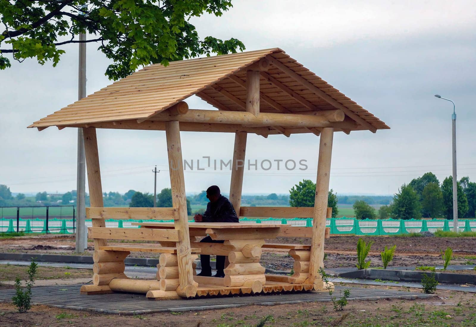 A person sits at a wooden picnic table in a gazebo, enjoying tranquility in a rural area with lush greenery.