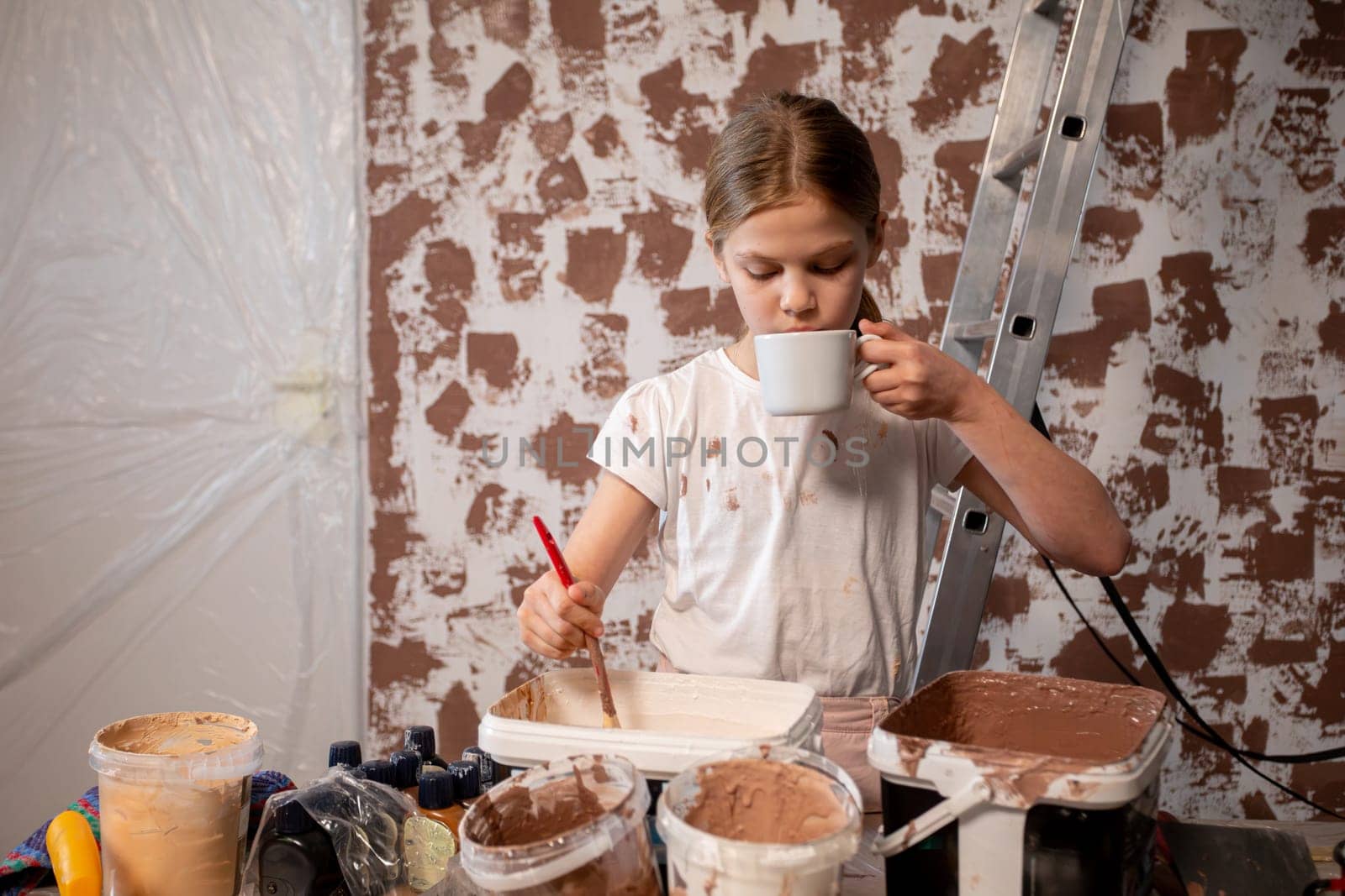 A young girl is creatively painting while holding a cup of her favorite drink in hand, showcasing her artistic talent