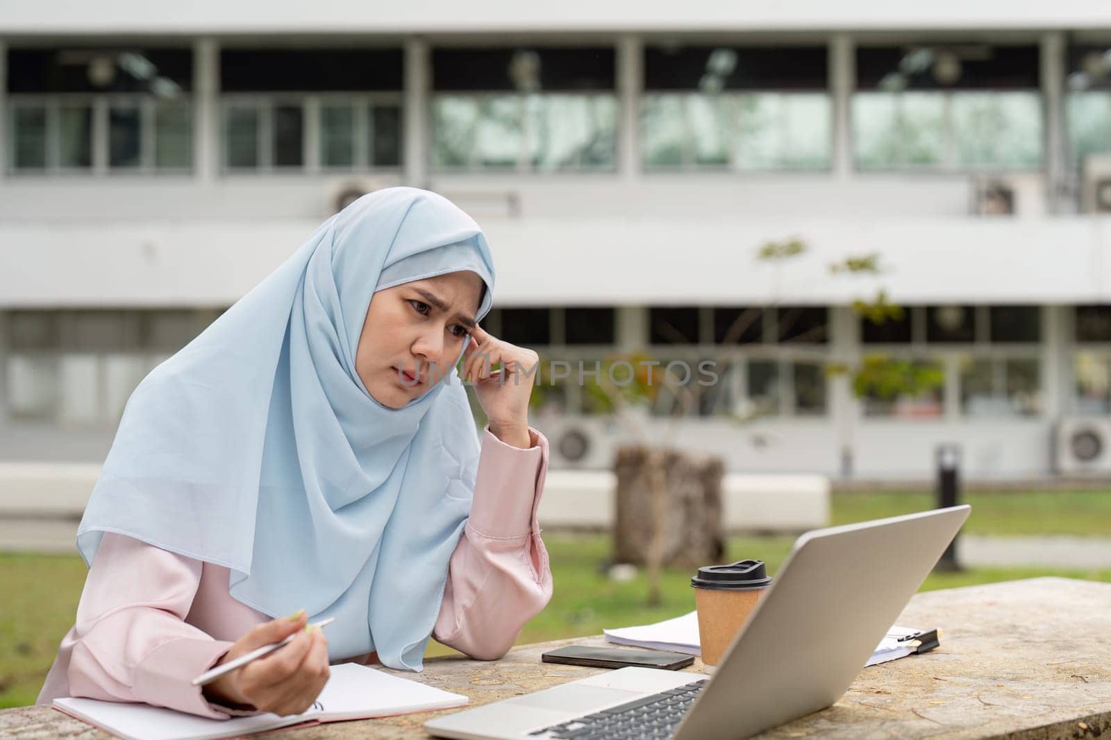 A Muslim woman in a hijab looks worried while working on her laptop outdoors, reflecting the challenges faced in her business activities.