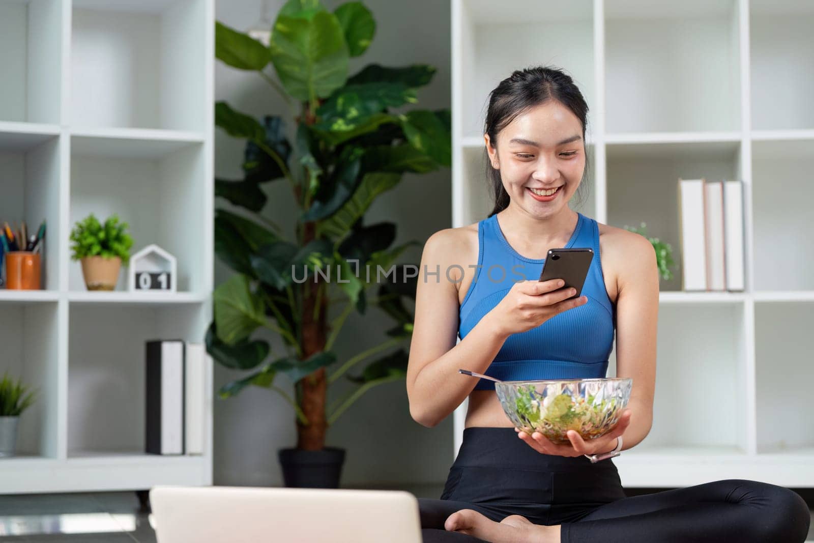 A woman sits cross-legged, holding a salad and smartphone, representing a modern approach to healthy living.