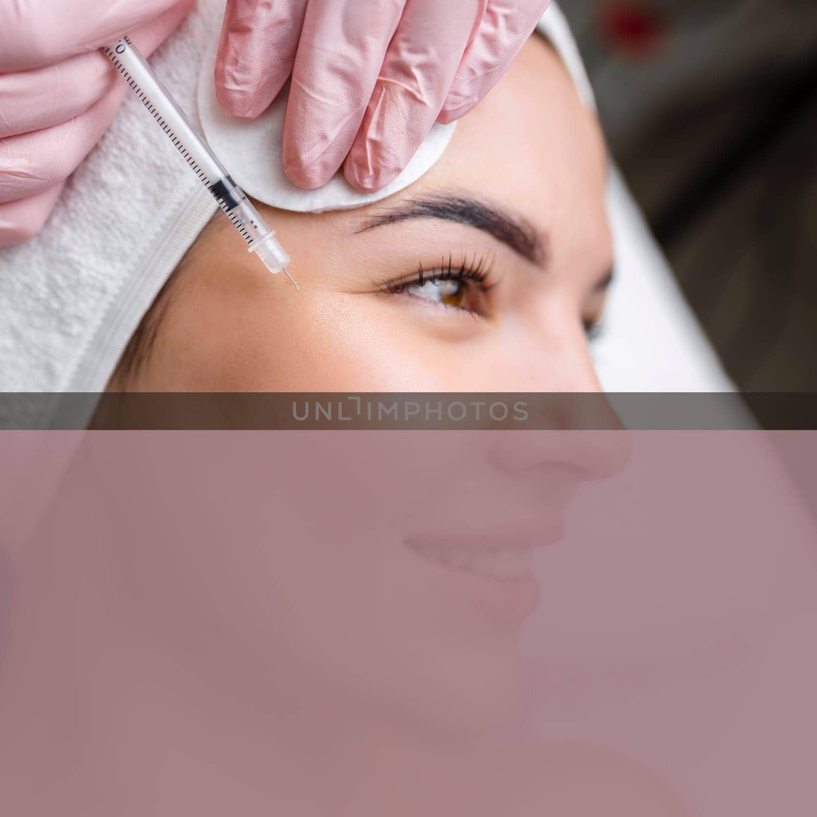 Close up of hands of young cosmetologist injecting botox in female face. She is standing and smiling. The woman is closed her eyes with relaxation.