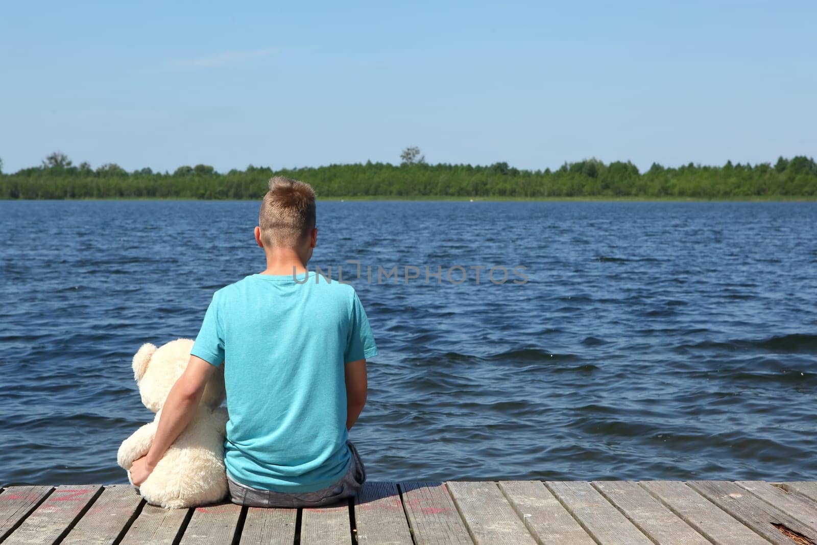 A young boy sits peacefully by the serene lake, holding his favorite teddy bear, fully enjoying natures tranquility