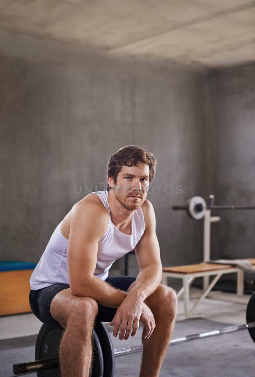 Serious young man looking at the camera while sitting on a weight in his private gym
