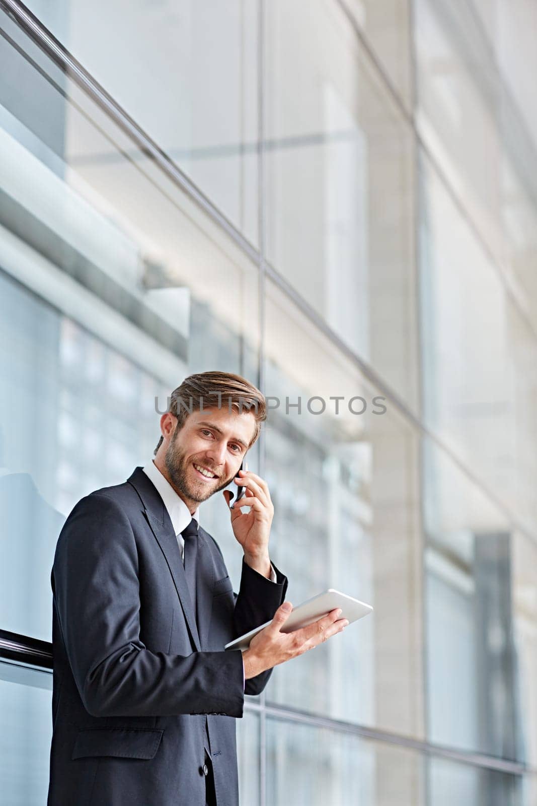 Corporate executive smiling at the camera while talking on his phone and holding at a digital tablet