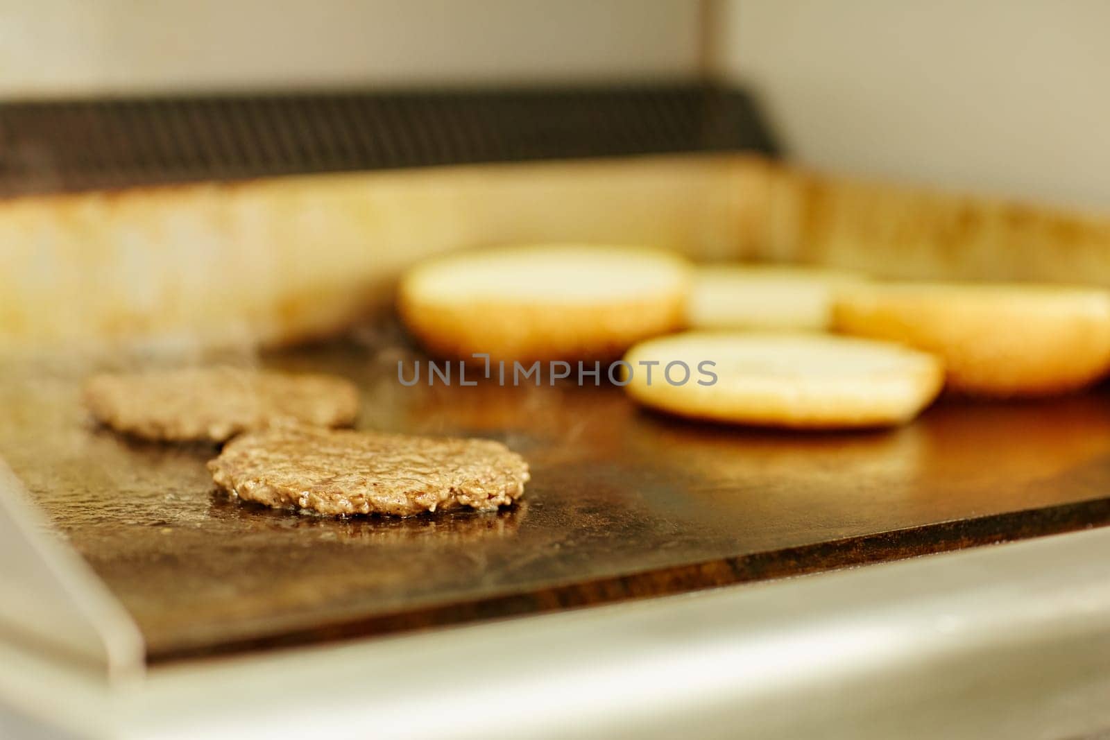 Two burger patties frying on a grill with hamburger buns toasting alongside.