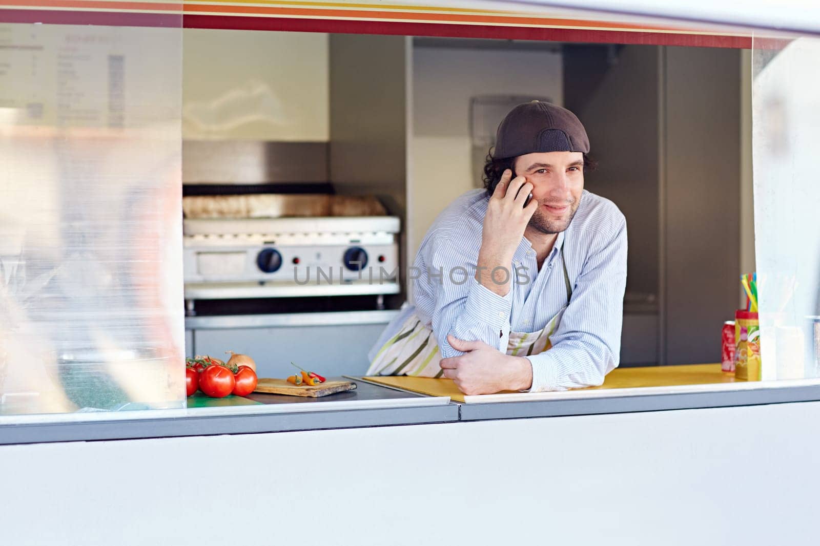 Male entrepeneur leaning on the counter and talking on the phone in his food stall where he makes and sells takeaway food