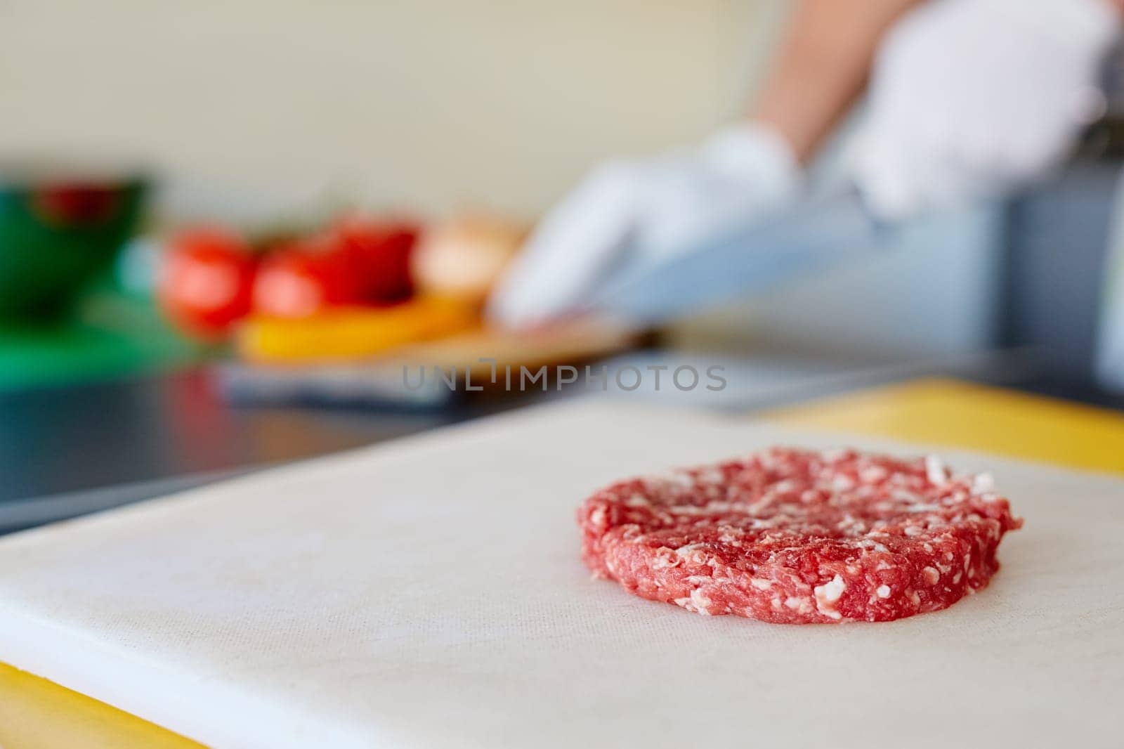 High quality raw hamburger patty sitting ready on a white board waiting for seasoning and cooking, while chef's hands prepare other ingredients in the background.