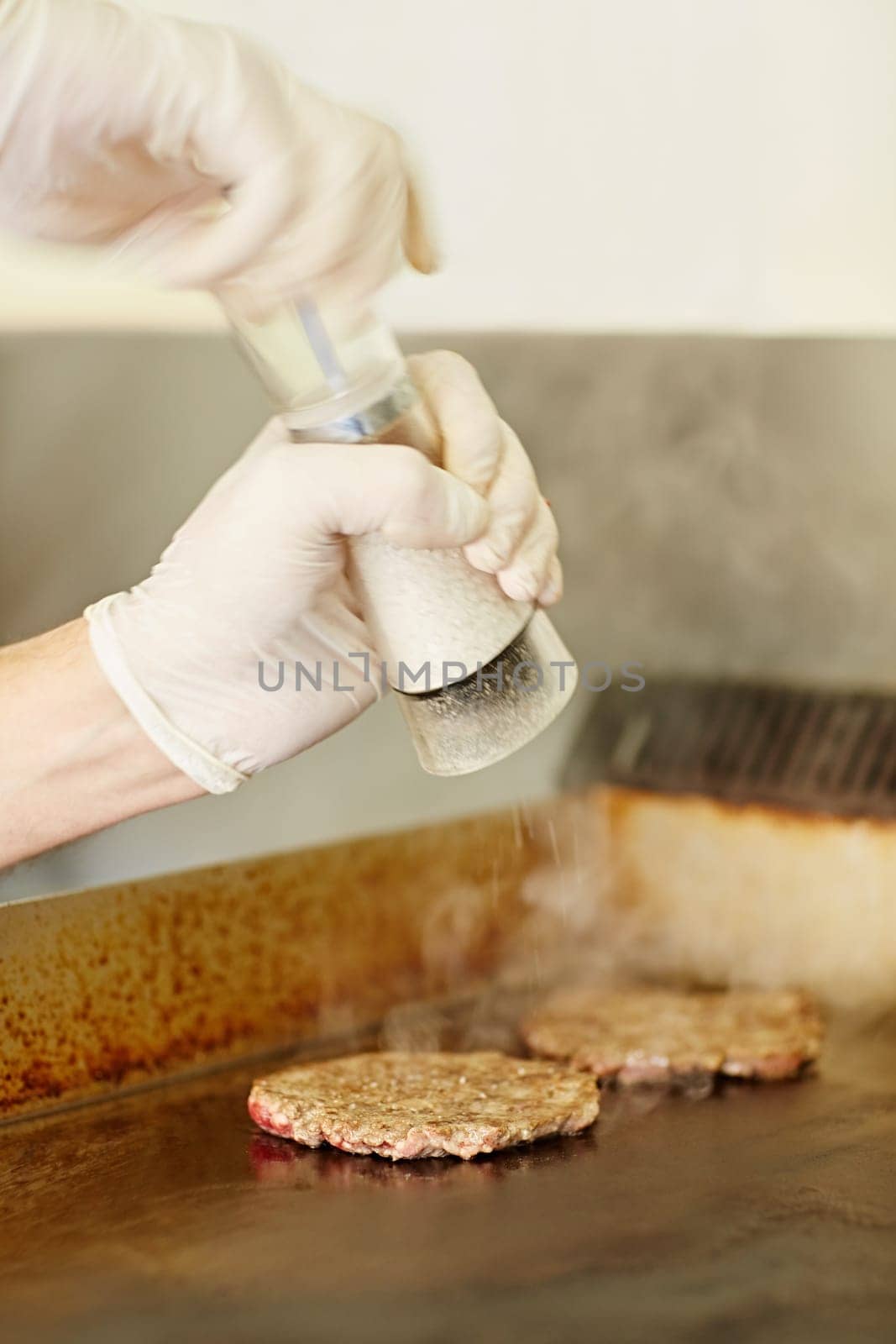 Cropped chef's hands wearing gloves busy grinding salt onto hamburger patties to season them while they are frying.