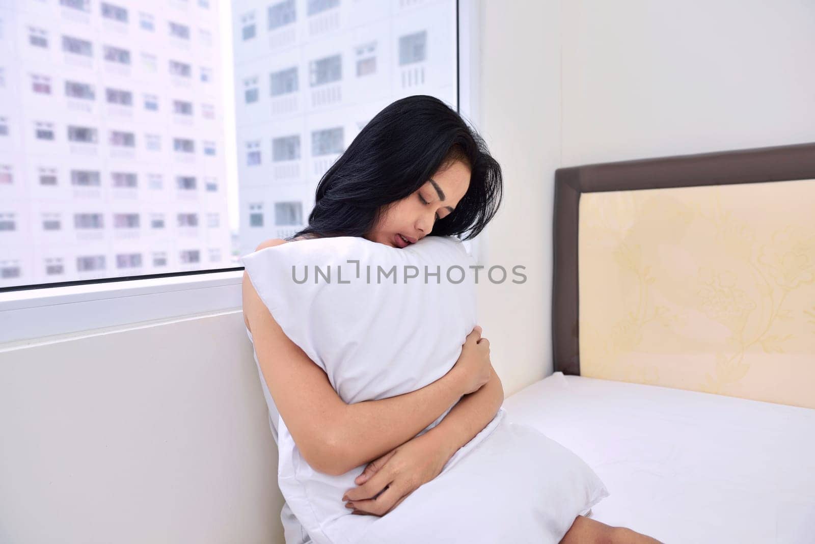 Beautiful asian woman sitting and hugging white pillow with window background