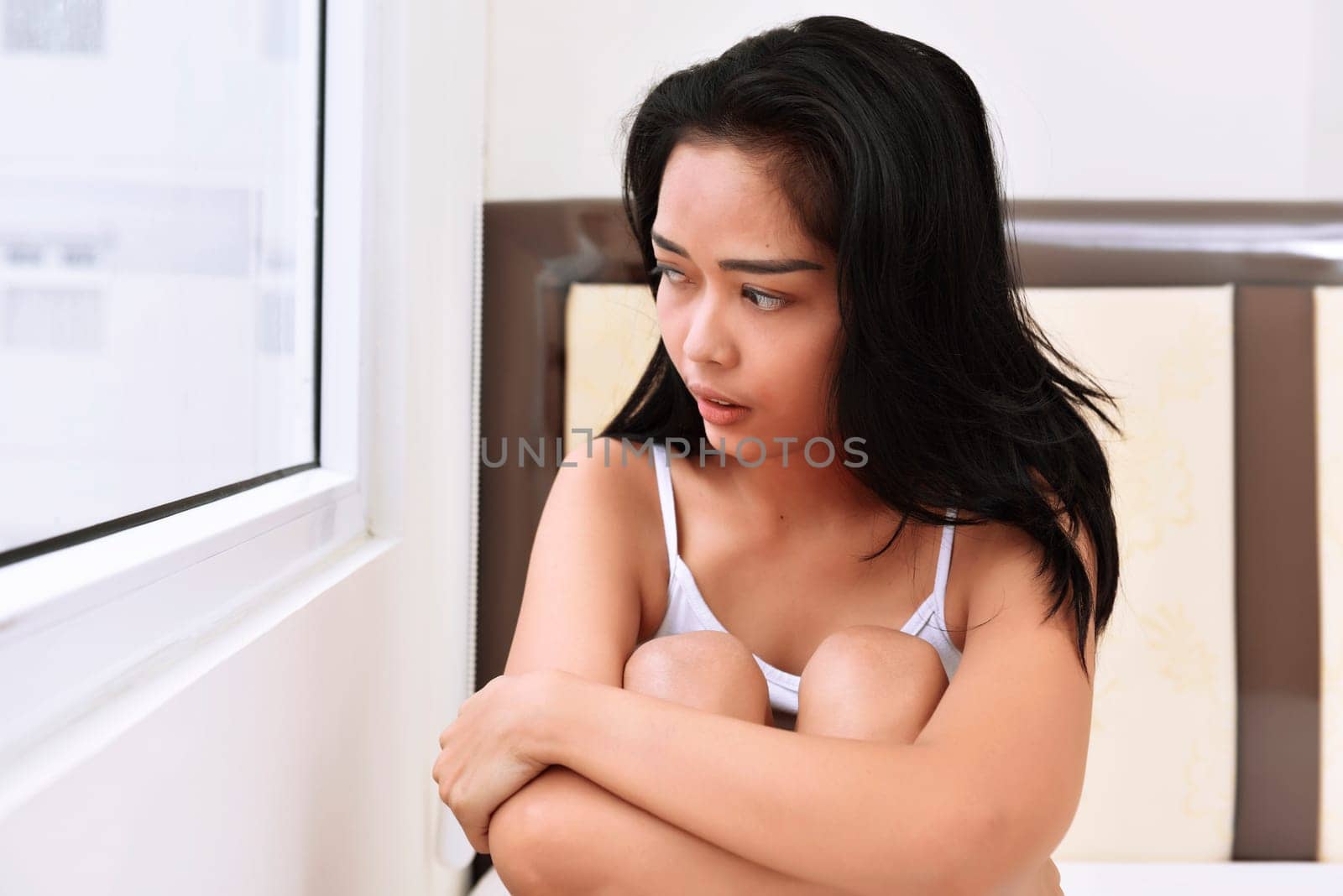 Beautiful asian girl in white shirt looking outdoors through the window from the bed