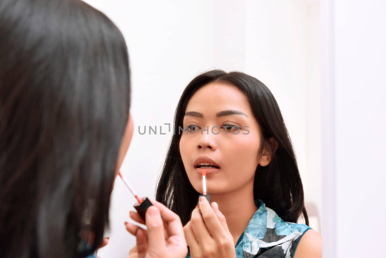 Beautiful asian girl applying lipstick while looking on the mirror at home