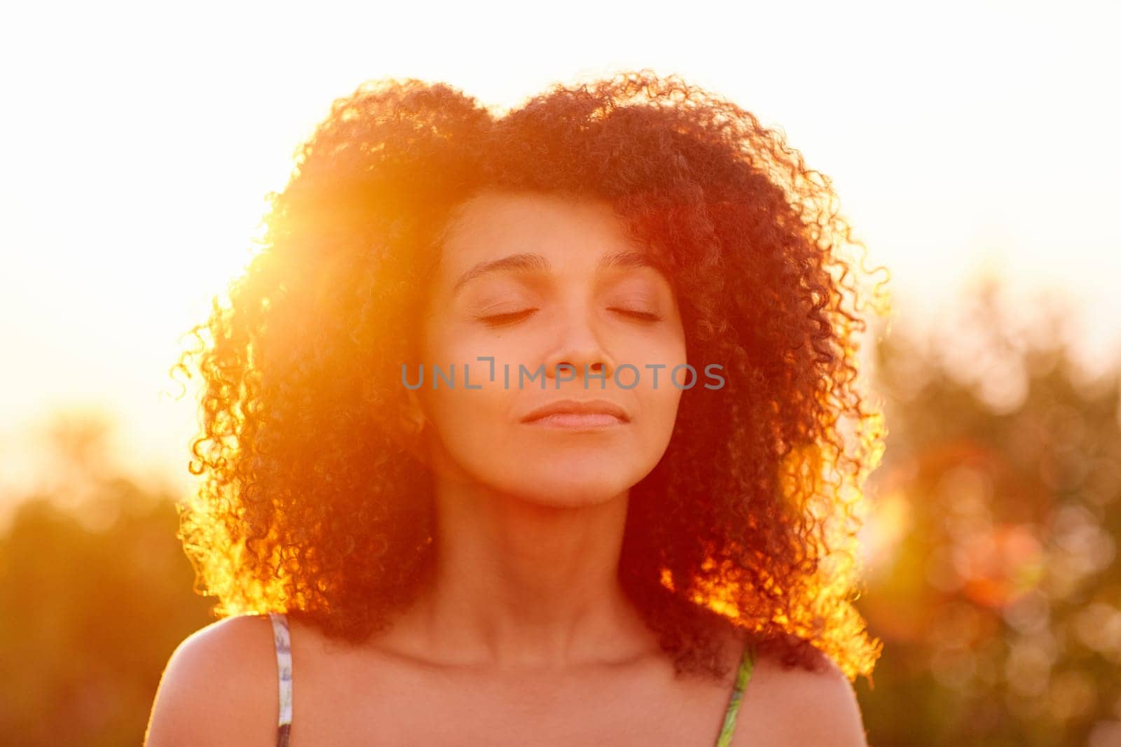 Beautiful woman with curly afro hair looking relaxed and happy with her eyes closed against a golden sunset with sun flare