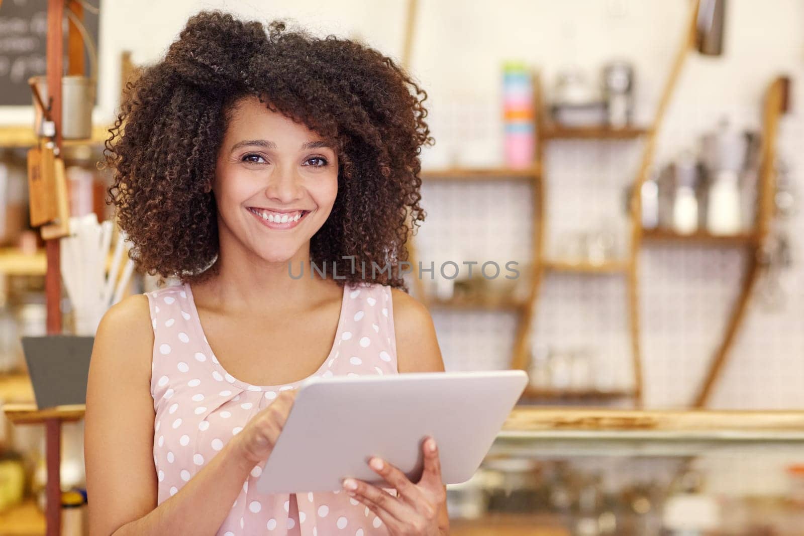 Portrait of a beautiful young woman with an afro hairstyle smiling at the camera while typing on her digital tablet in her coffee shop