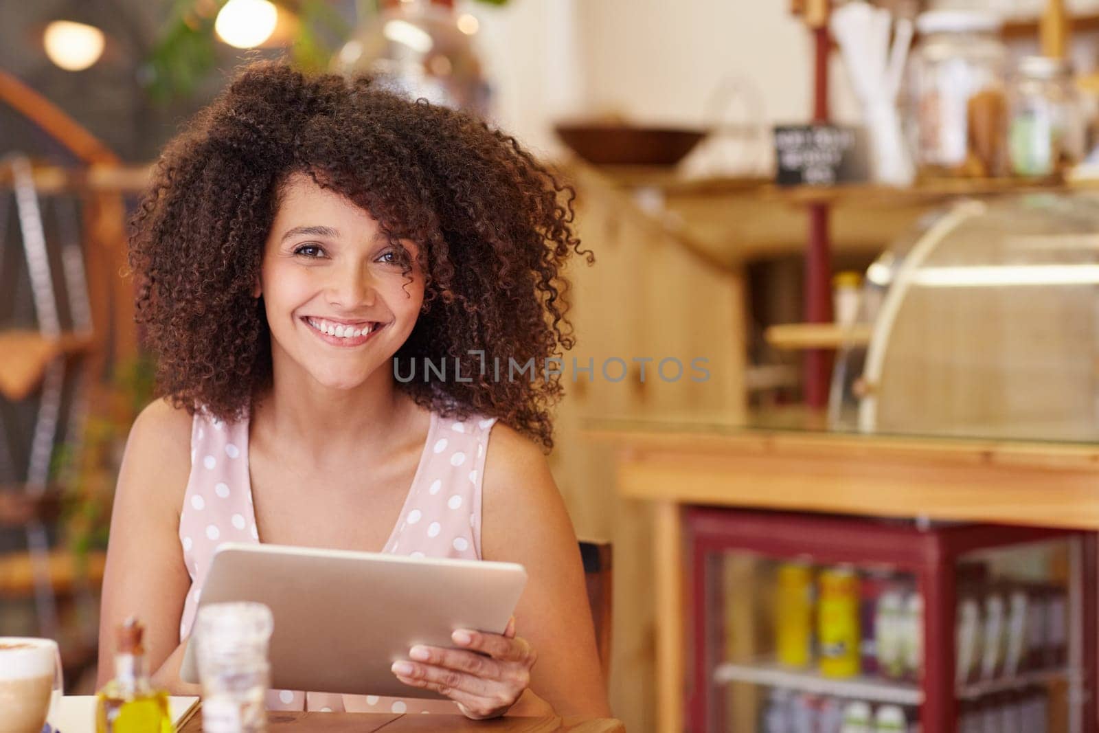 Beautiful young mixed race woman smiling at the camera confidently while using her digital tablet in a coffee shop