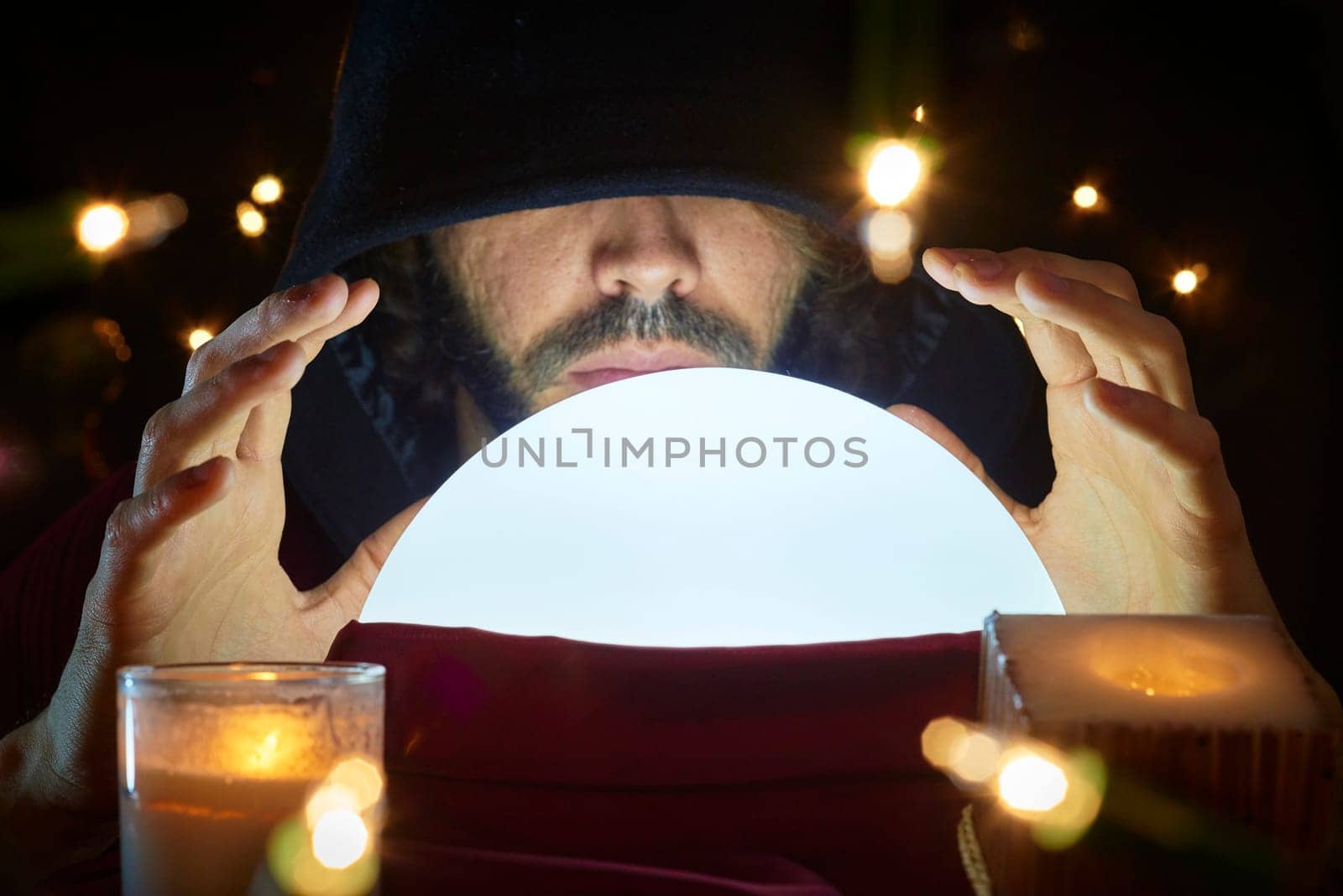 Very low key portrait of hooded man with eyes covered reading fortune on bright crystall ball, surrounded by candle light.