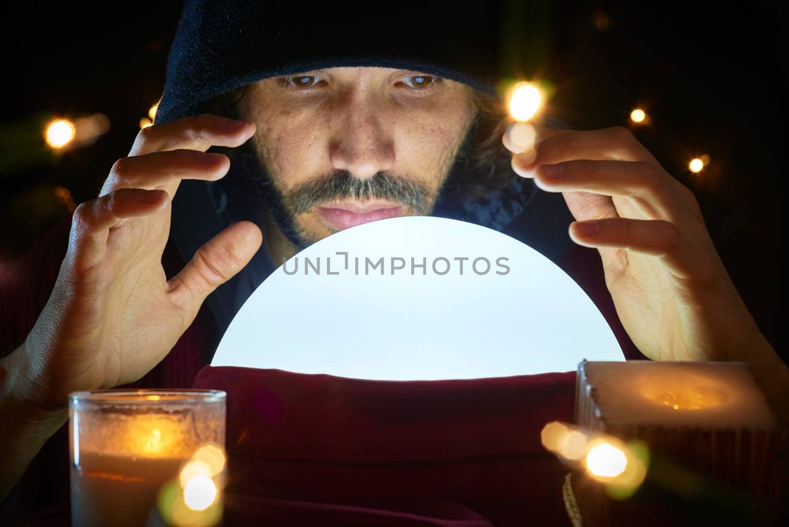 Very low key portrait of hooded man reading fortune on bright crystall ball, surrounded by candle light.
