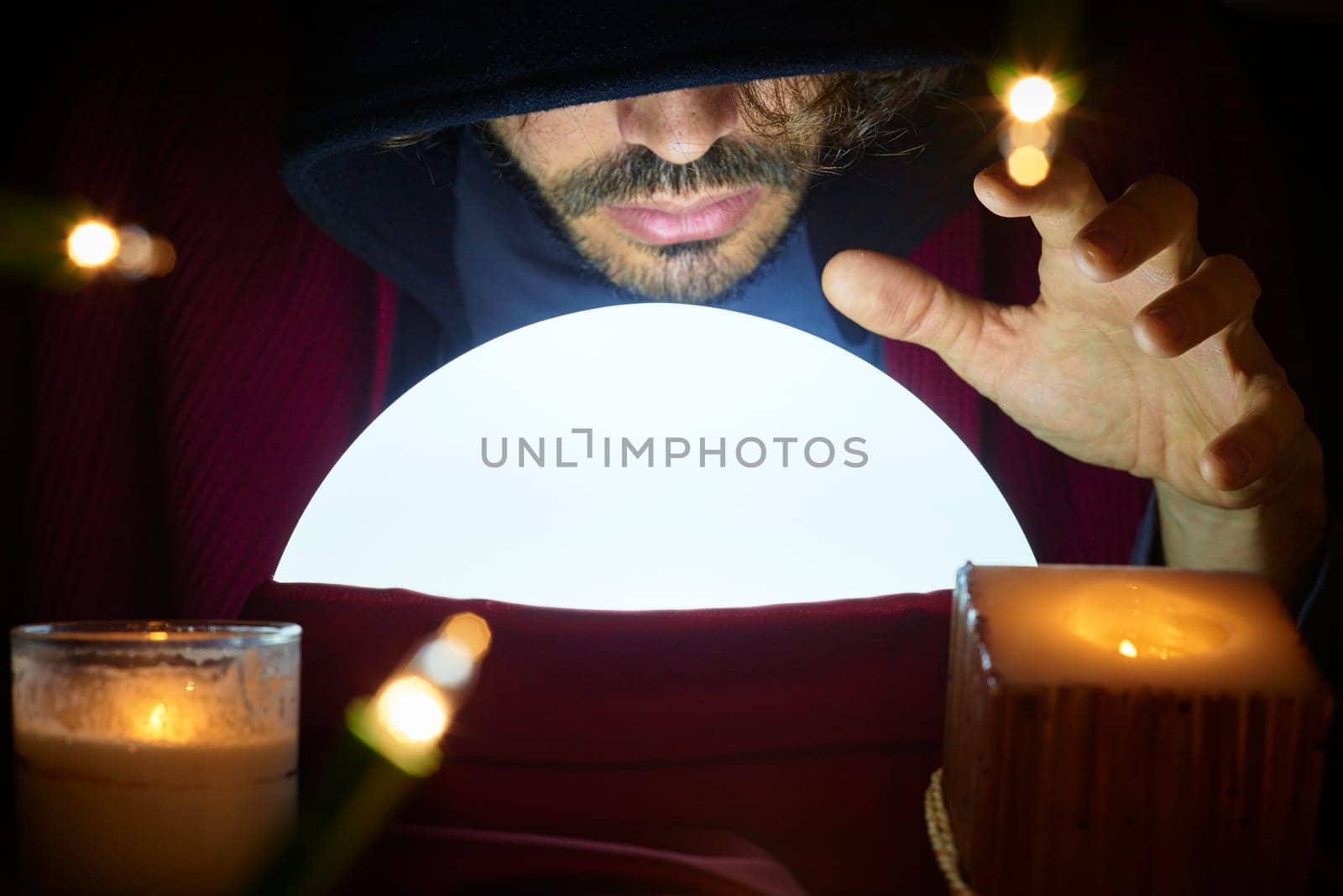 Very low key portrait of hooded man with eyes covered reading fortune on bright crystall ball, surrounded by candle light.