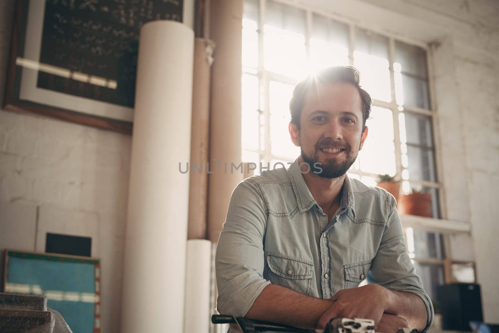 Portrait of a handsome young male designer looking positively at the camera with gentle sunflare coming in through the window of his studio
