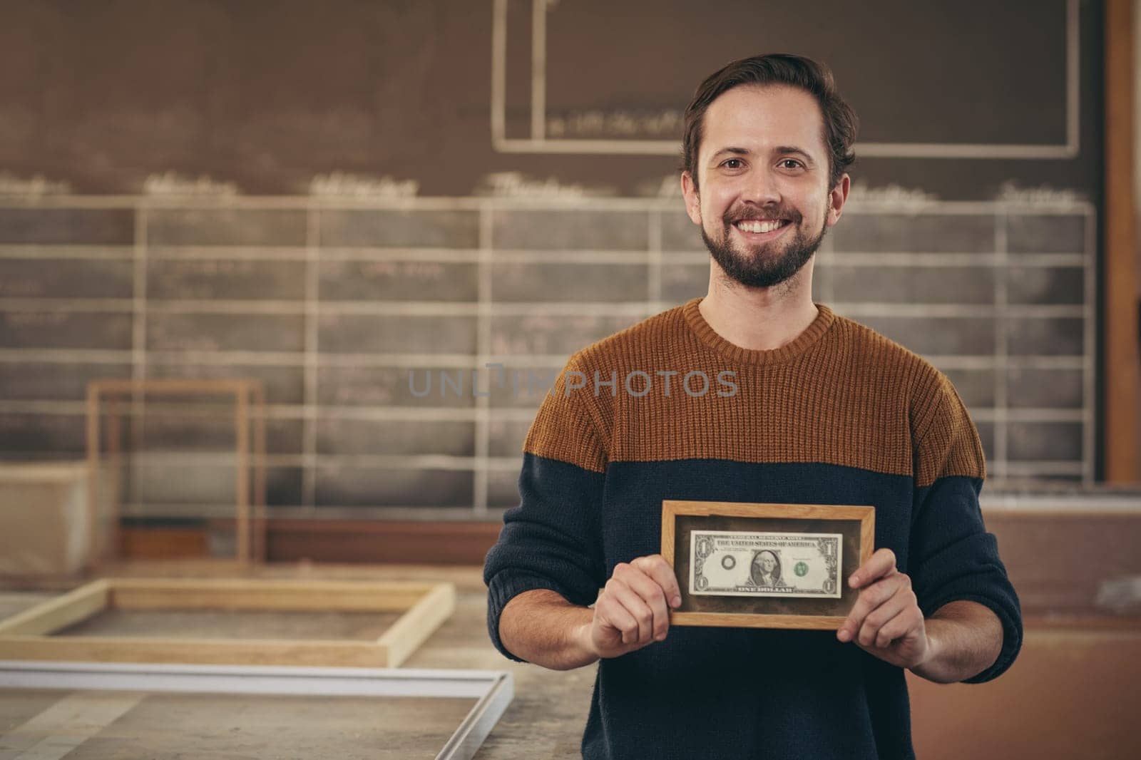 Smiling craftsman entrepreneur standing proudly in his workshop and displaying a bank note that has been carefully framed