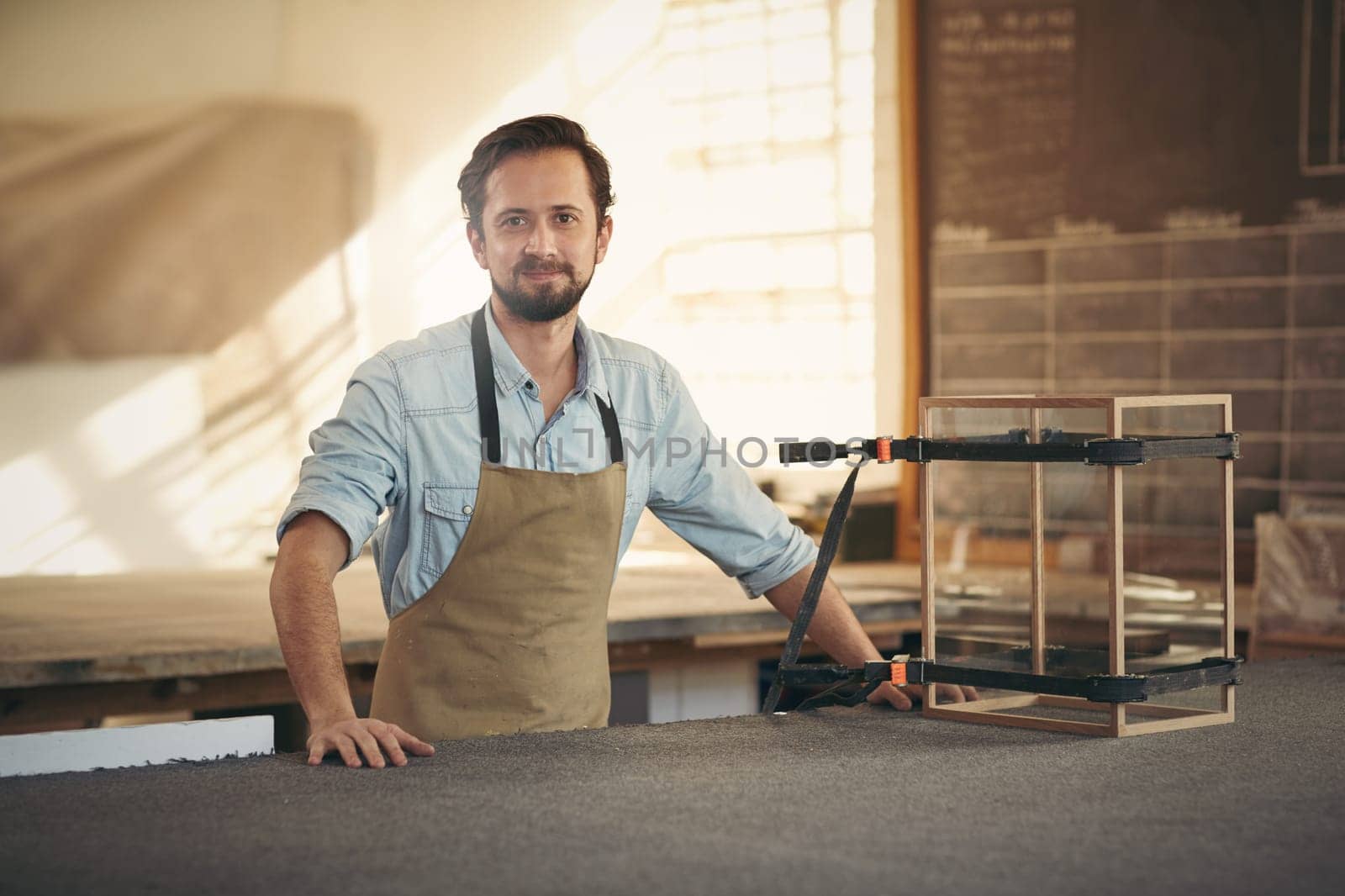 Craftsman smiling at the camera while standing proudly in his workshop alongside a wood and glass display case that he is making with care