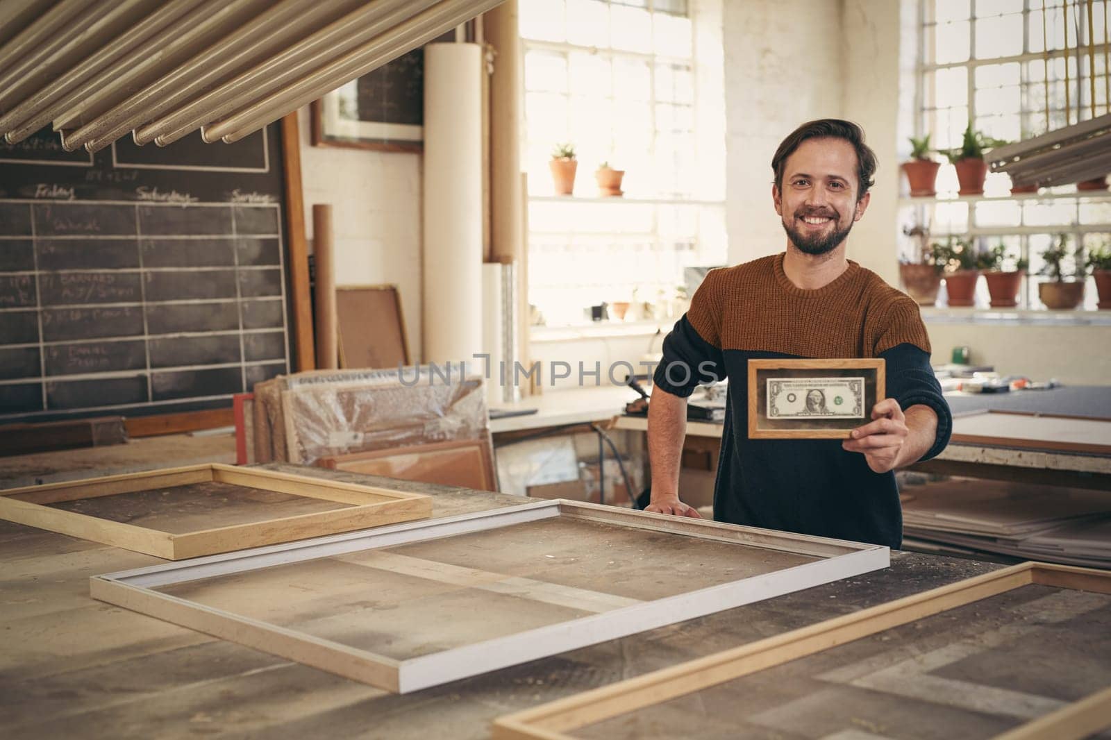 Young entrepreneur smiling proudly in his workshop while holding up a framed bank note