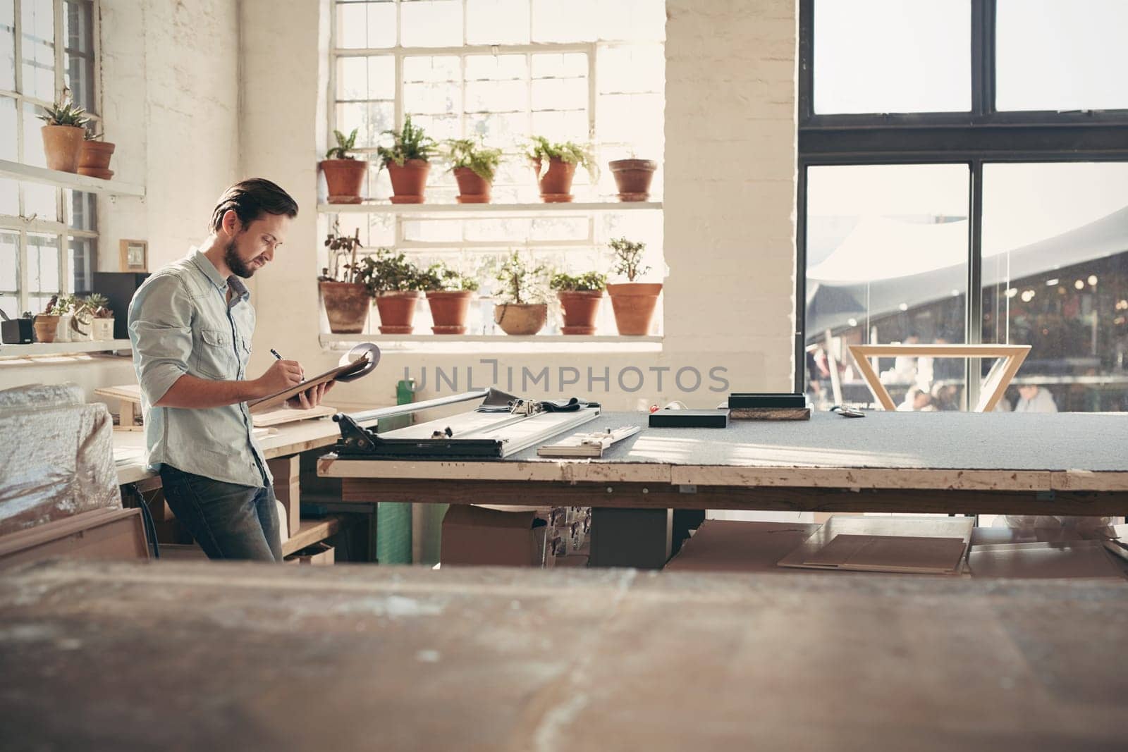 Young male business owner standing in his studio workshop checking figures and stock on his clipboard on a sunny afternoon