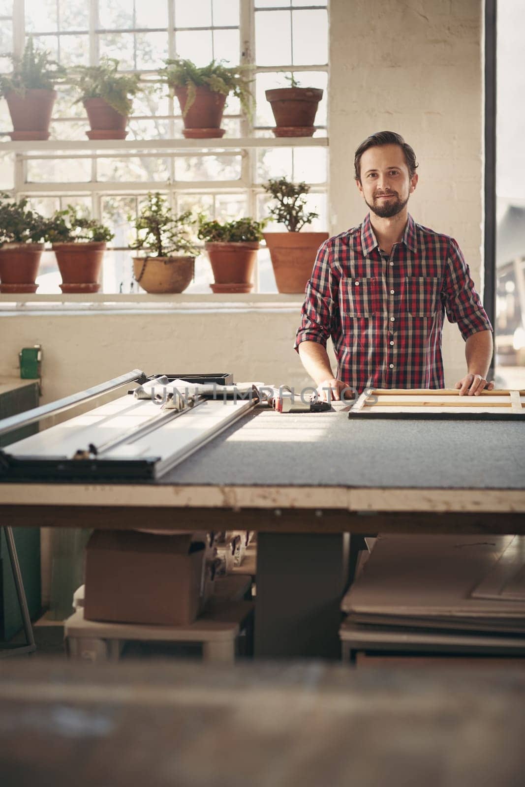 Young male designer entrepreneur standing proudly in his studio at his workbench smiling with beautiful light coming in through the window