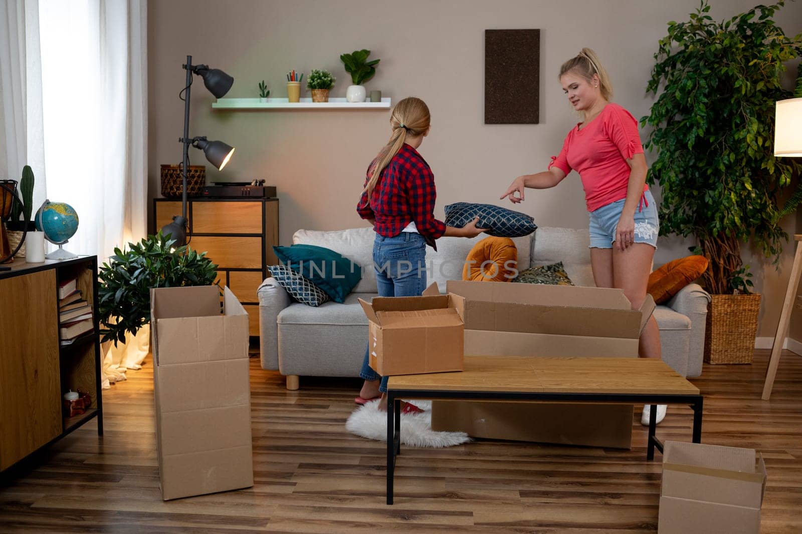 Two friends unpack boxes in a stylish living room filled with plants and furniture, creating a vibrant vibe