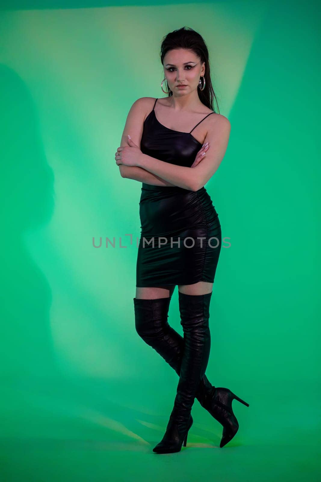 A stylish model poses confidently in a black dress and thigh-high boots on a vibrant green backdrop.