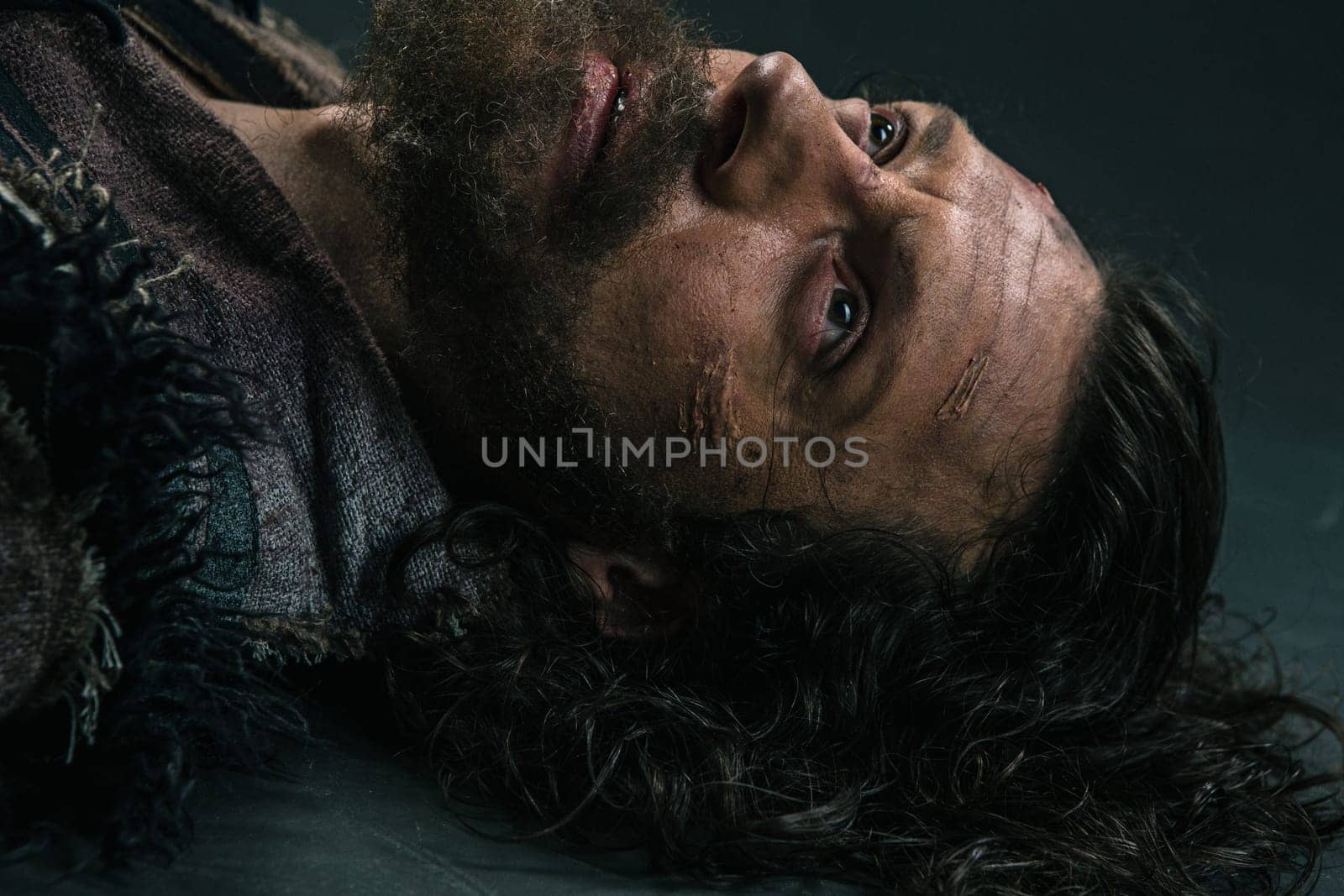 Halloween zombie lying on paving asphalt. Vampire hipster with red blood outdoors. Holiday celebration concept. Man with bloody beard and hair. War soldier with wounds on face.