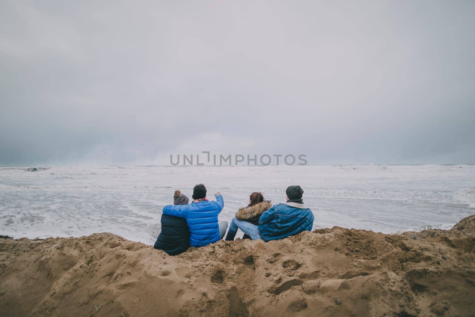 Four teenagers sit facing away from the camera whilst admiring the views on a beach in winter.