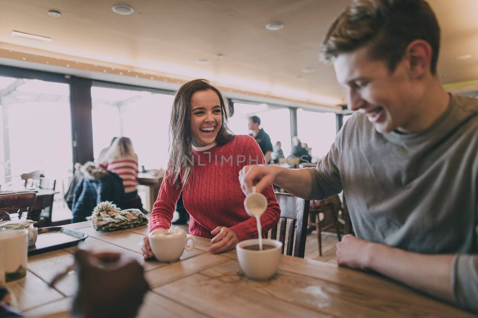 A young couple laugh in a cafe in winter whilst the male pours milk into a cup of coffee.