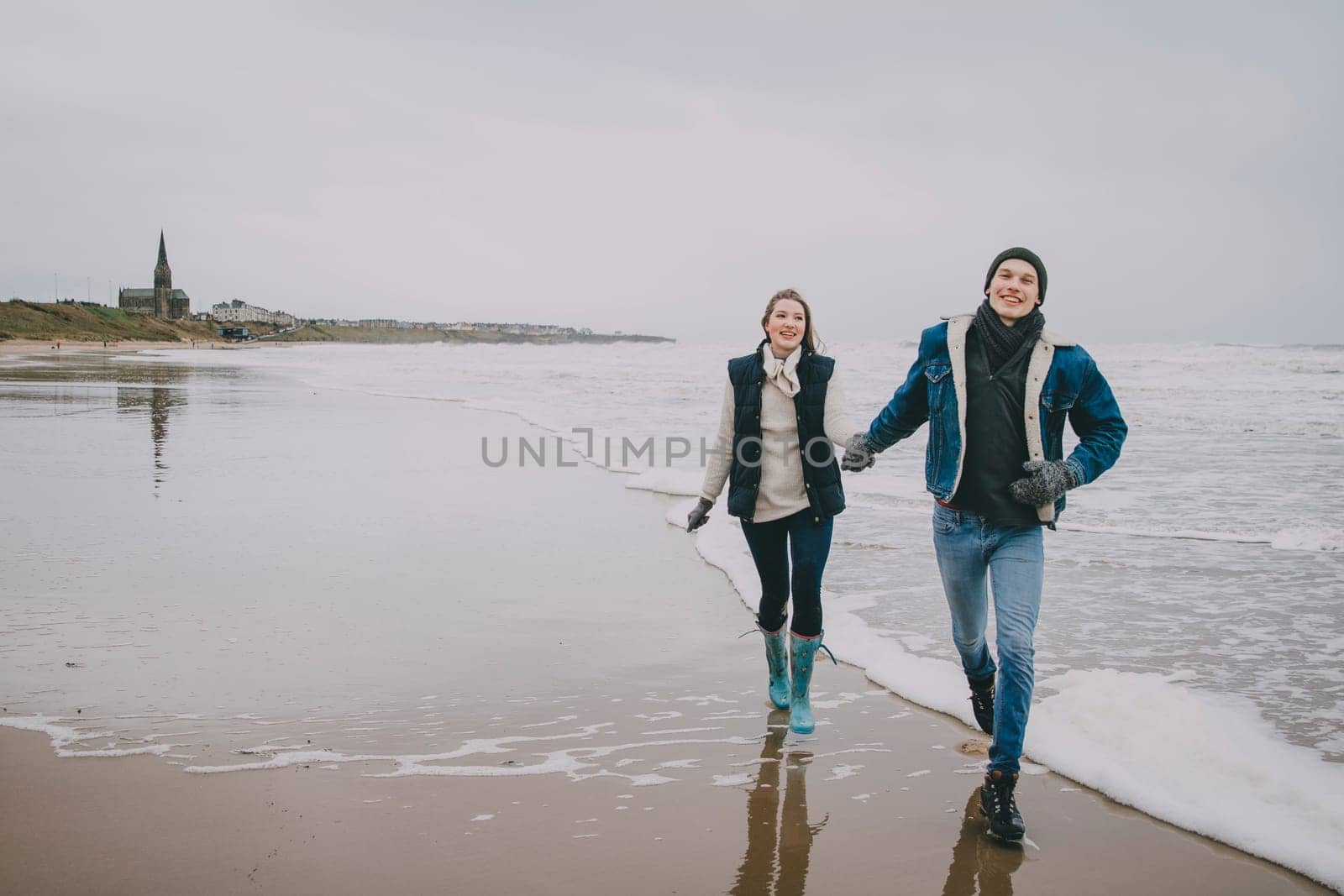 A young couple smile as they walk along a winter beach together.