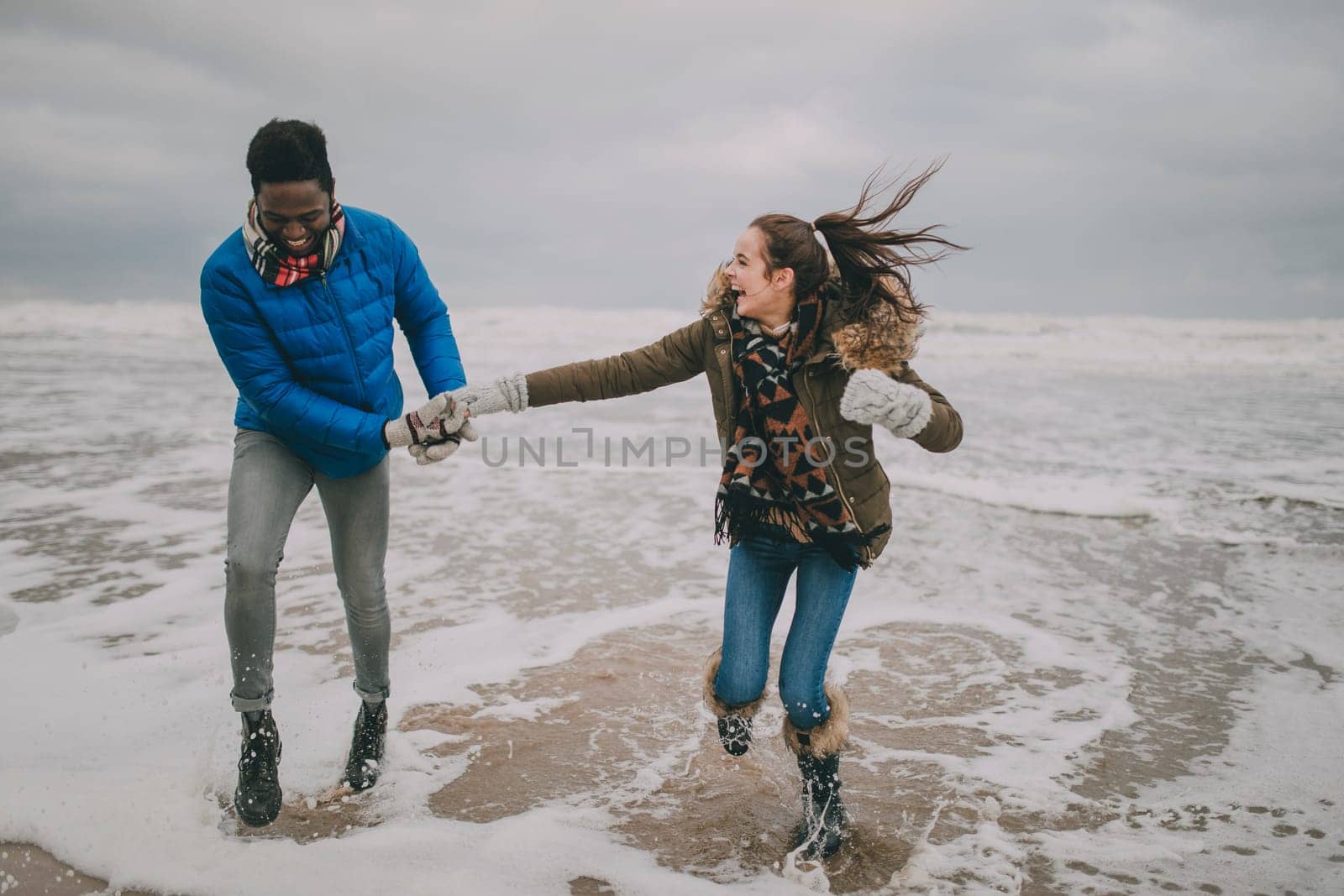 A young couple laugh, hold hands and paddle in the sea on Tynemouth beach in winter.