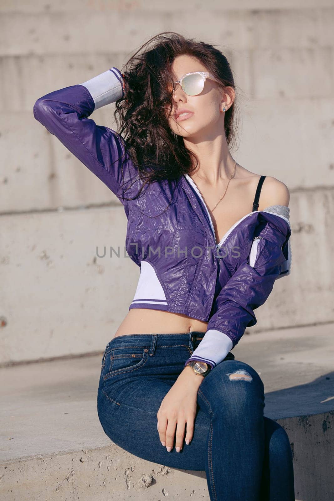 Fashionable young woman in sunglasses, lilac short windbreaker and jeans. Sits on a concrete structure, bare shoulder. Women's fashion. Urban way of life.