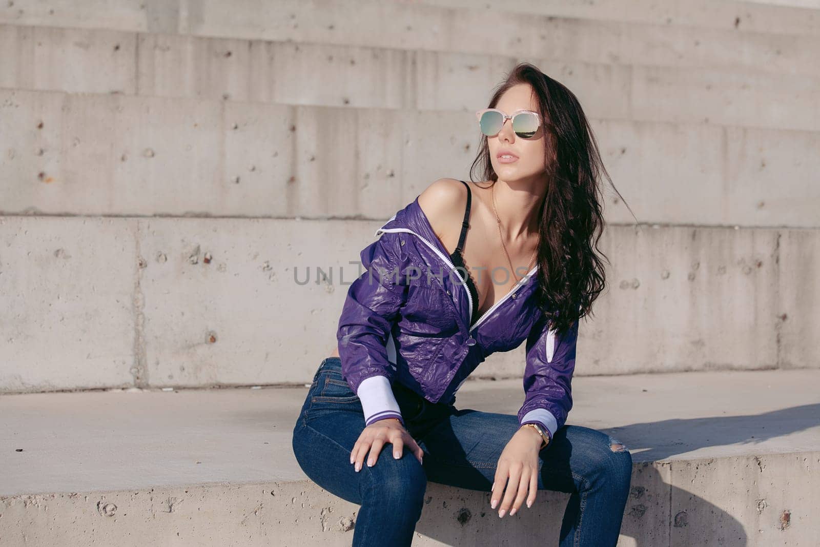 Fashionable young woman in sunglasses, lilac short windbreaker and jeans. Sits on a concrete structure, bare shoulder. Women's fashion. Urban way of life.