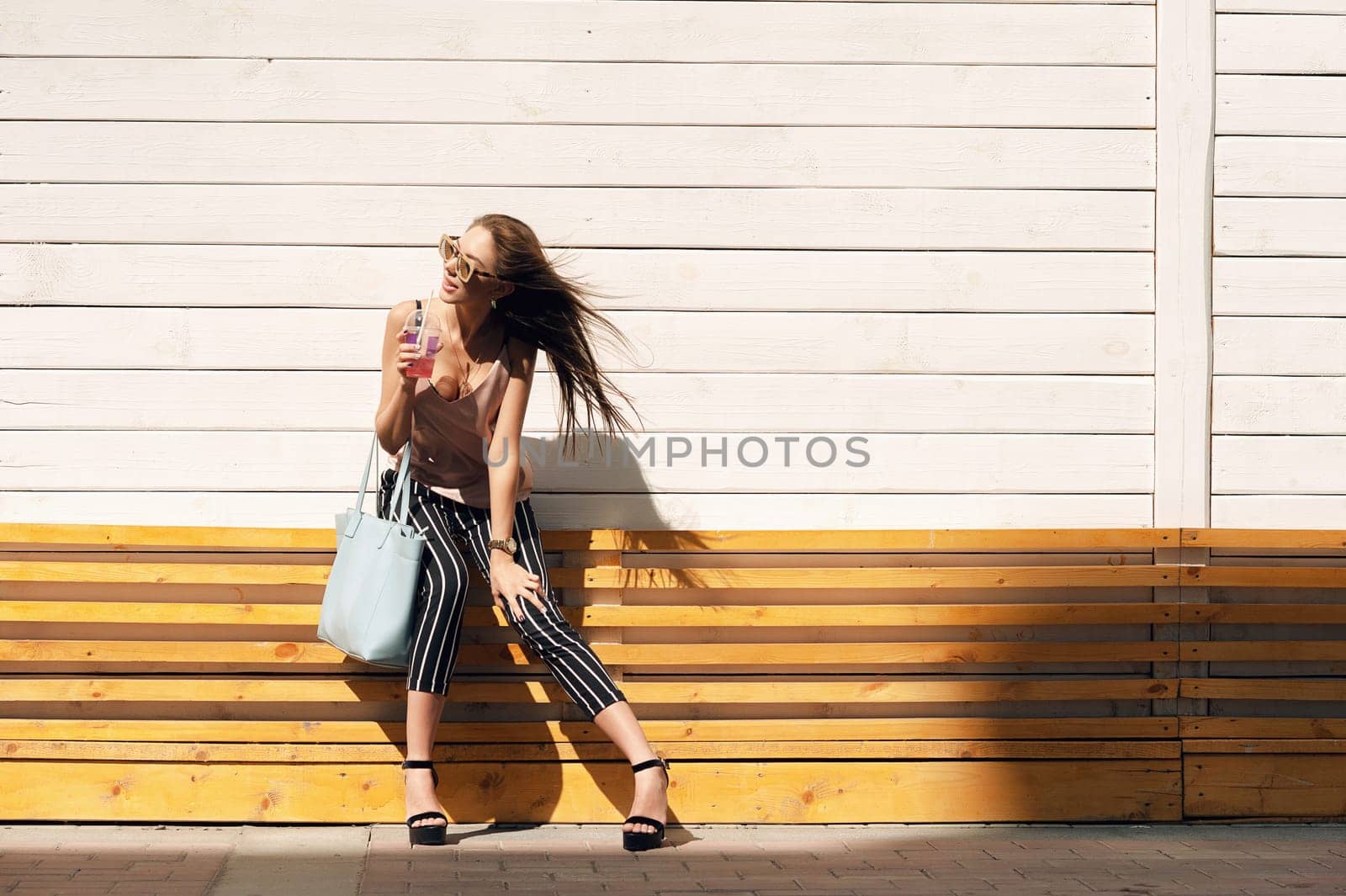 Outdoor portrait of a young beautiful fashionable happy lady wearing sunglasses, in a wooden frame, against a wooden, white wall. The model wears stylish clothes, a beige blouse, black breeches in a white stripe, a bag of light blue. The girl looks away and drinks pink mors and tubes. Women's fashion. Urban way of life.